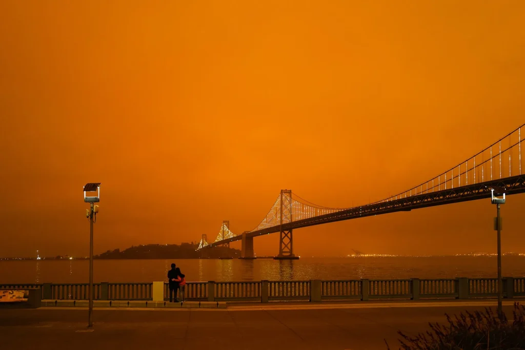 A suspension bridge and waterfront under a dark orange smoke-filled sky